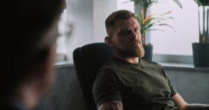 Depressed military man in camouflage with PTSD sits in chair, shares feelings with psychotherapist or psychologist on rehabilitation therapy session. Post traumatic stress disorder.