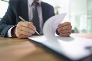 Confident male employee flips page of contract signing after reading. Man in classic suit puts signature on paper sheet making important decision in company.