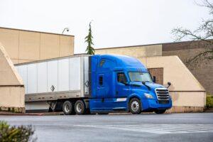 Industrial grade blue big rig long haul semi truck with extended cab for truck driver rest and dry van semi trailer unloading delivered cargo standing in store dock gate at raining time.